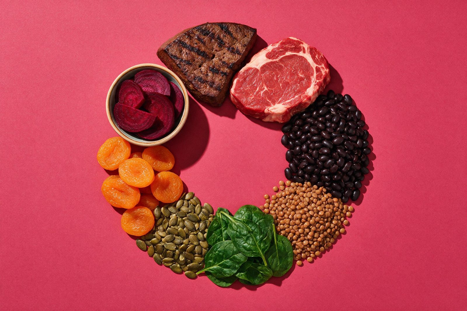 Alimentos ricos em ferro — fígado, carne vermelha magra, feijão preto, lentilha, espinafre, sementes de abóbora, damasco seco e beterraba — sobre fundo rosa