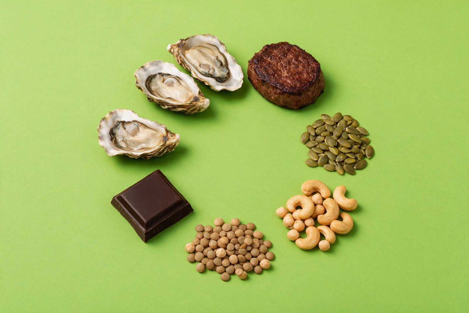 Alimentos ricos em zinco — ostras, carne bovina, sementes de abóbora, castanhas, grão-de-bico, chocolate amargo e lentilha — sobre fundo verde-limão