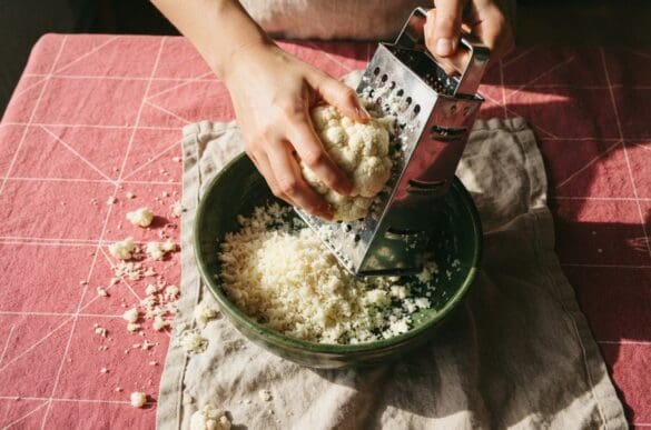 Mãos ralando couve-flor sobre uma tigela verde já com arroz de couve-flor, sobre toalha rosa da Frutifica