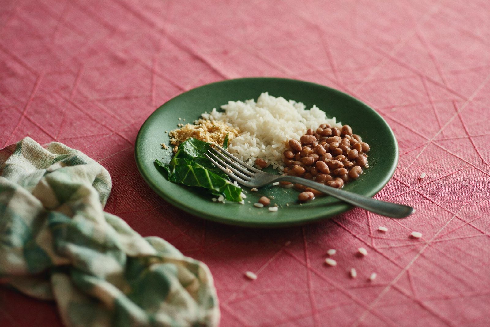 Prato verde com arroz branco, feijão, couve refogada e farofa sobre toalha rosa da Frutifica