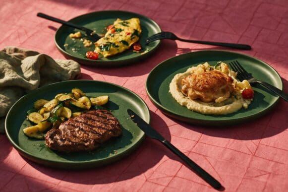 Cardápio low carb em três pratos verdes: bife com abobrinha, frango com purê de couve-flor e omelete com espinafre, sobre toalha rosa
