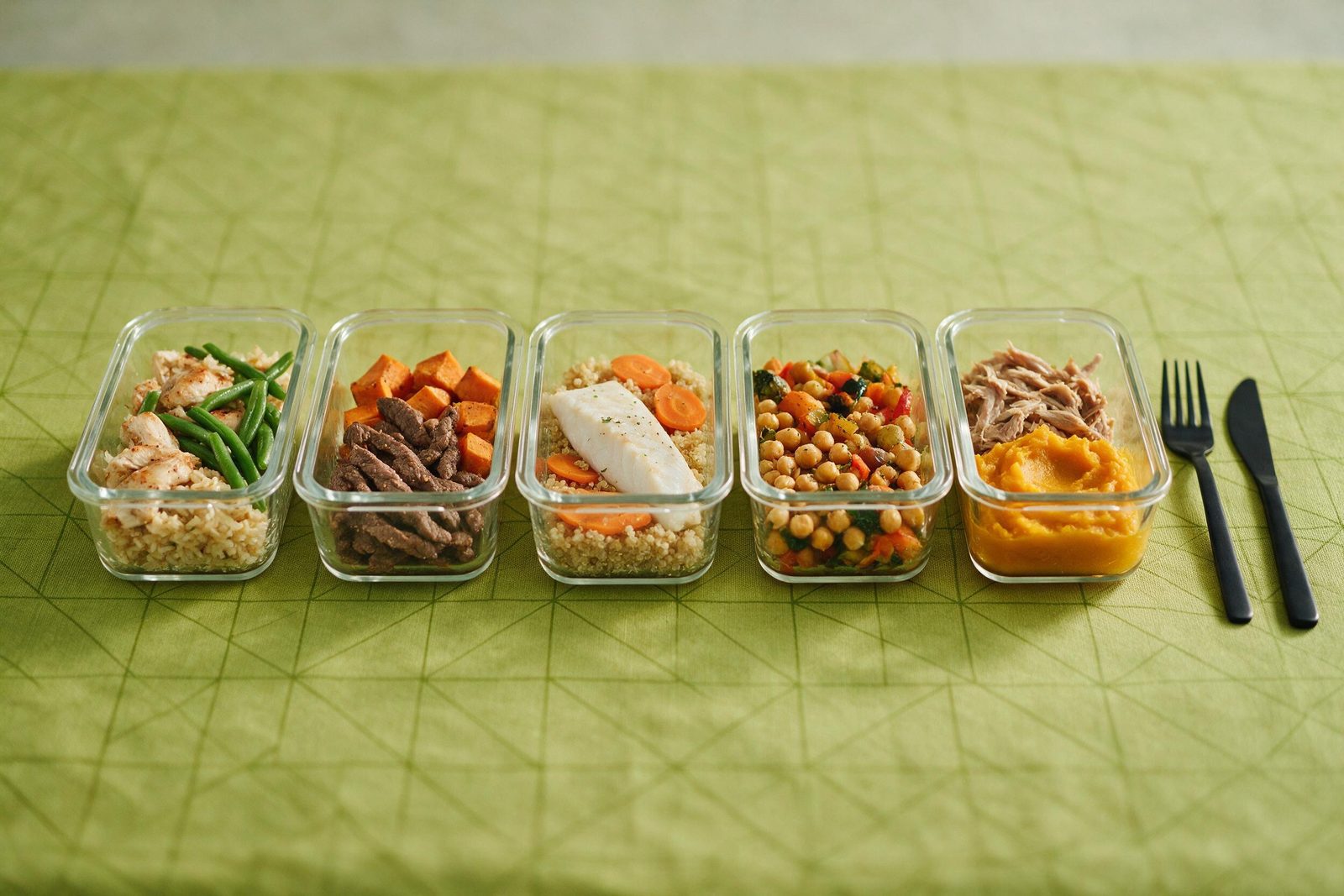 Cinco potes de vidro com refeições variadas de proteína, grão e legumes alinhados sobre toalha verde-limão