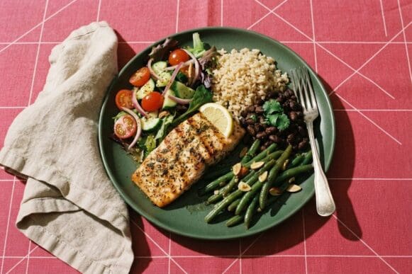 Prato verde com peixe grelhado, arroz integral, feijão preto e legumes, indicado para quem controla a glicemia, sobre toalha rosa