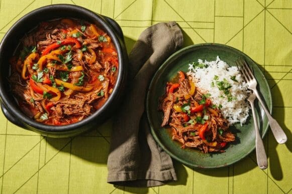 Panela de barro com carne louca desfiada com pimentões e cebola, com porção servida em prato verde e arroz, sobre toalha verde-limão