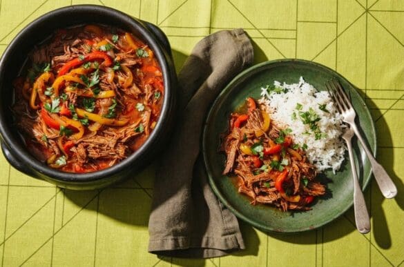 Panela de barro com carne louca desfiada com pimentões e cebola, com porção servida em prato verde e arroz, sobre toalha verde-limão