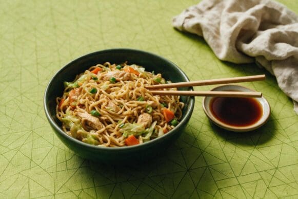 Tigela verde-escura de yakisoba com frango e legumes, hashi apoiado na borda e molho shoyu, sobre toalha verde-limão