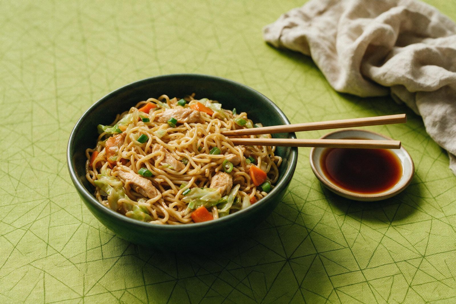 Tigela verde-escura de yakisoba com frango e legumes, hashi apoiado na borda e molho shoyu, sobre toalha verde-limão