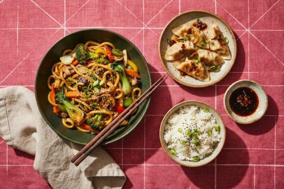 Pratos orientais — macarrão salteado com legumes, guioza e arroz branco — com hashi, sobre toalha rosa