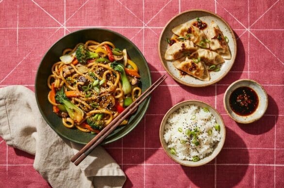 Pratos orientais — macarrão salteado com legumes, guioza e arroz branco — com hashi, sobre toalha rosa