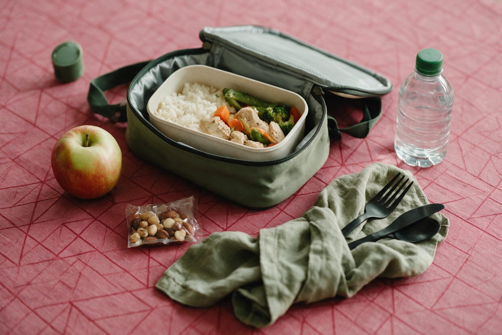 Bolsa térmica aberta com marmita de arroz, frango e legumes, maçã, garrafa de água e castanhas, pronta para levar, sobre toalha rosa