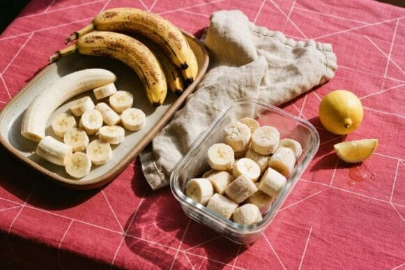 Bananas inteiras e em rodelas em bandeja para congelar, com uma porção já congelada em pote, sobre toalha rosa