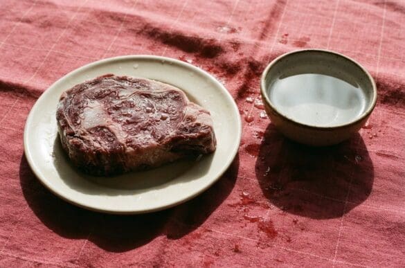 Peça de carne bovina descongelando em prato ao lado de uma tigela de água fria, sobre toalha rosa da Frutifica