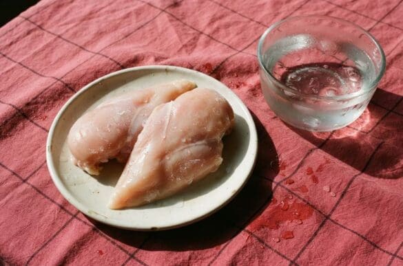 Peitos de frango descongelando em prato ao lado de uma tigela de água fria, sobre toalha rosa da Frutifica