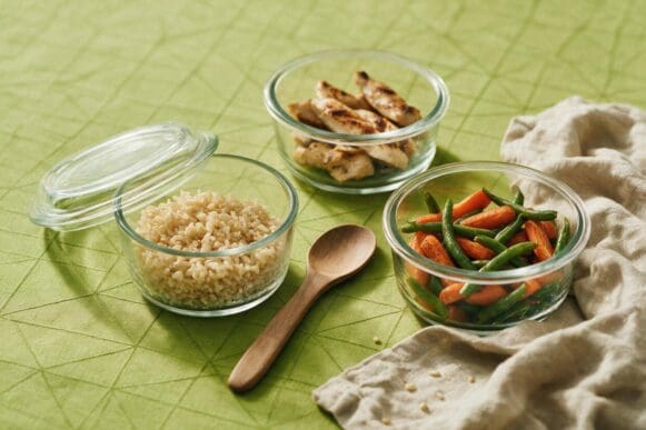 Potes de vidro sendo montados com arroz integral, frango grelhado e legumes sobre toalha verde-limão da Frutifica