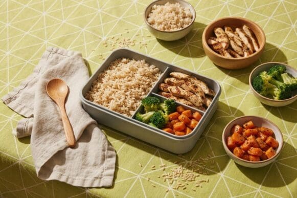 Marmita fit sendo montada com arroz integral, frango e legumes, com os ingredientes em tigelas ao redor, sobre toalha verde-limão