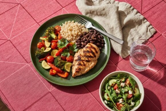 Prato verde com porções equilibradas de frango grelhado, arroz integral, feijão e bastante legume, sobre toalha rosa