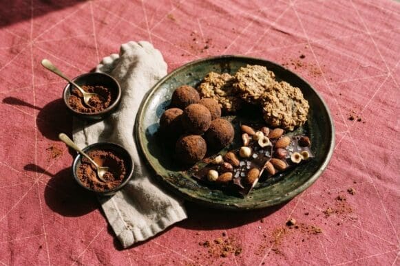 Doces fit — brigadeiros de cacau, cookies de aveia e banana e barras com castanhas — em prato verde sobre toalha rosa