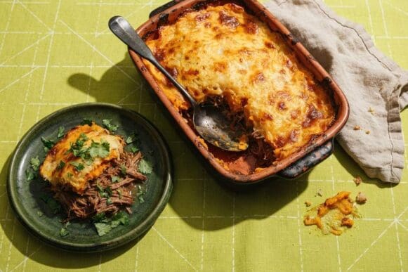 Travessa de escondidinho de batata-doce gratinado com carne desfiada, com uma porção servida em prato verde, sobre toalha verde-limão