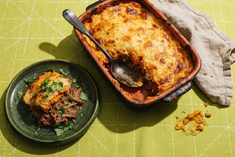Travessa de escondidinho de batata-doce gratinado com carne desfiada, com uma porção servida em prato verde, sobre toalha verde-limão