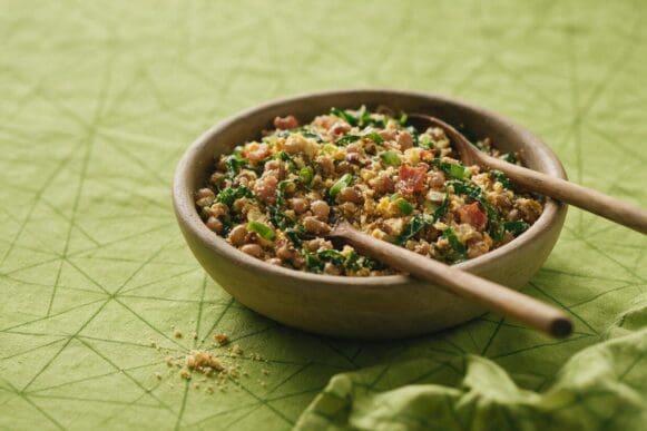 Tigela de barro com feijão tropeiro — feijão, farofa, bacon, couve e ovo — colher de pau apoiada, sobre toalha verde-limão