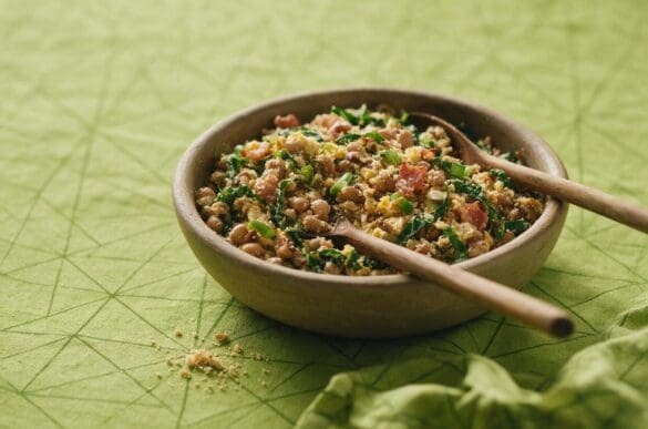 Tigela de barro com feijão tropeiro — feijão, farofa, bacon, couve e ovo — colher de pau apoiada, sobre toalha verde-limão