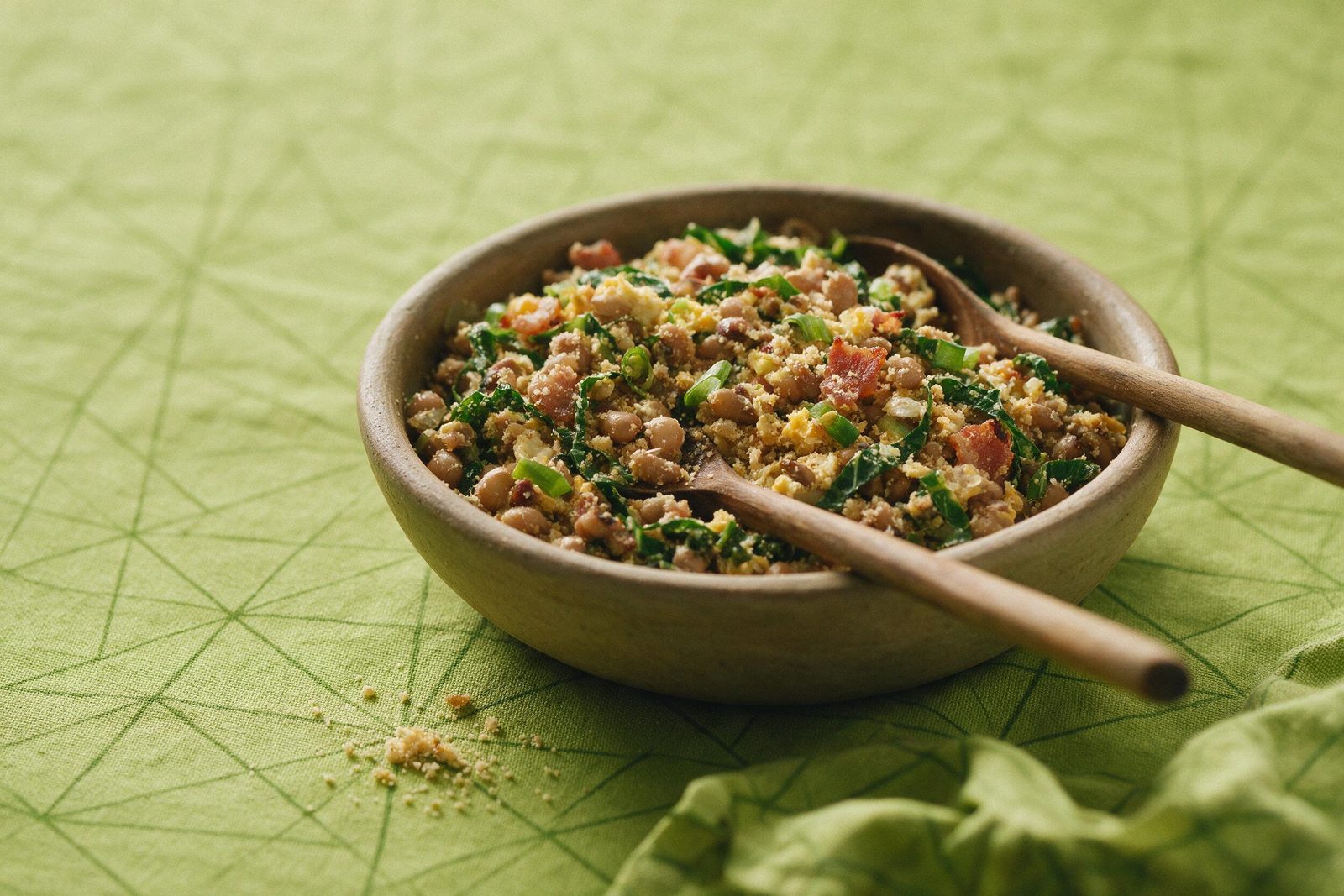 Tigela de barro com feijão tropeiro — feijão, farofa, bacon, couve e ovo — colher de pau apoiada, sobre toalha verde-limão
