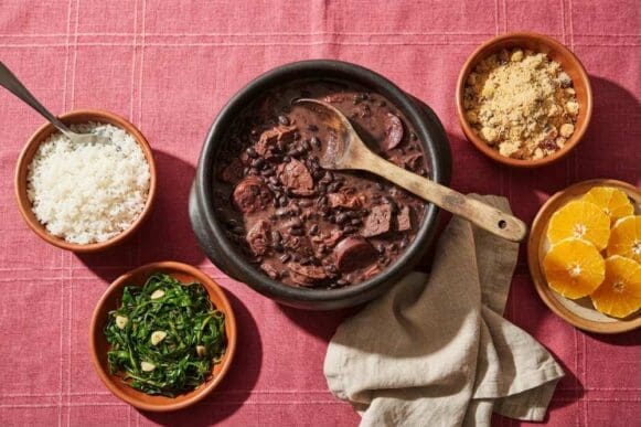 Panela de barro com feijoada leve, arroz, couve refogada, farofa e laranja em tigelas ao redor, sobre toalha rosa