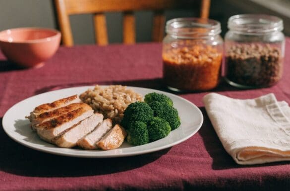 prato porcionado com frango grelhado, arroz integral e brócolis sobre mesa de casa, ao lado de potes de vidro com comida organizada em porções