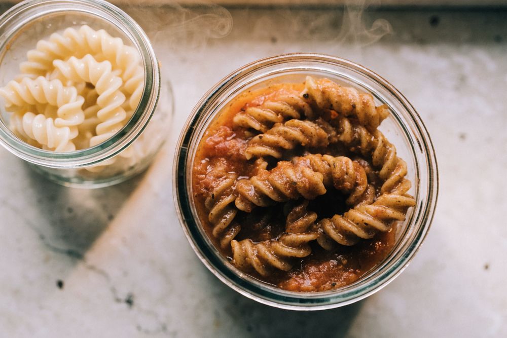 macarrão integral al dente porcionado com molho em pote de vidro ao lado de macarrão comum, pronto para congelar