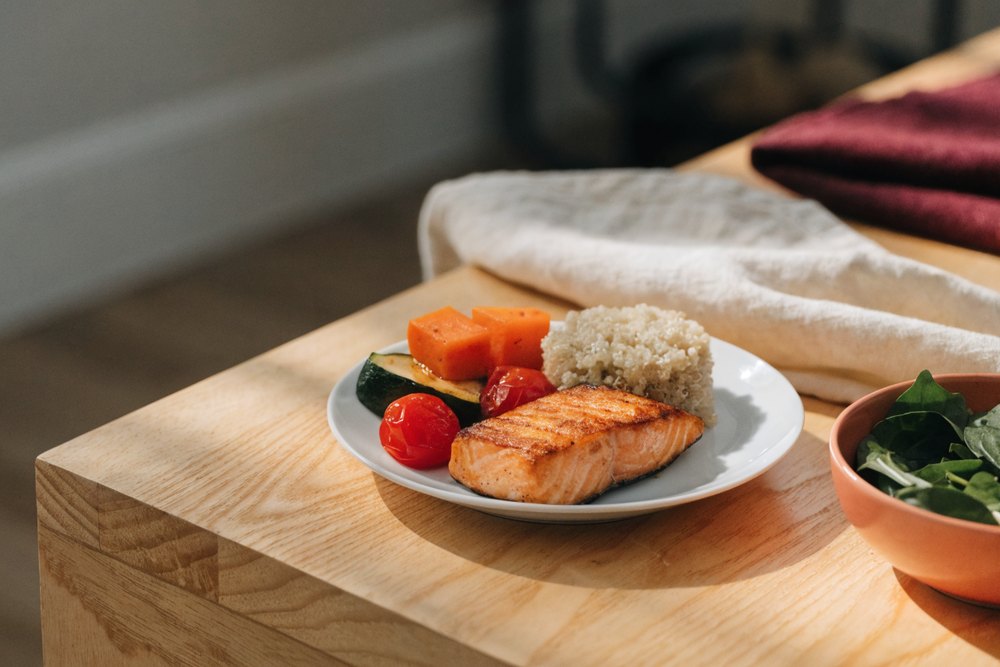 prato pequeno e bem porcionado com salmão grelhado, quinoa e legumes coloridos assados sobre mesa de madeira clara em casa