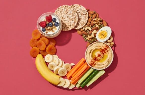 Lanches saudáveis — iogurte com frutas vermelhas, bolacha de arroz, castanhas, ovo cozido, palitos de cenoura e pepino com homus e banana — sobre fundo rosa