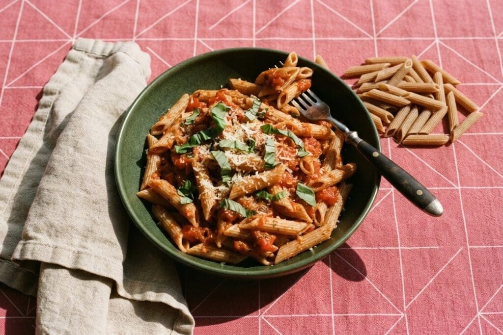Tigela verde de macarrão integral ao sugo com manjericão e queijo ralado ao lado de penne cru, sobre toalha rosa