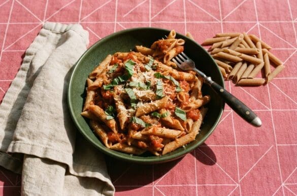 Tigela verde de macarrão integral ao sugo com manjericão e queijo ralado ao lado de penne cru, sobre toalha rosa