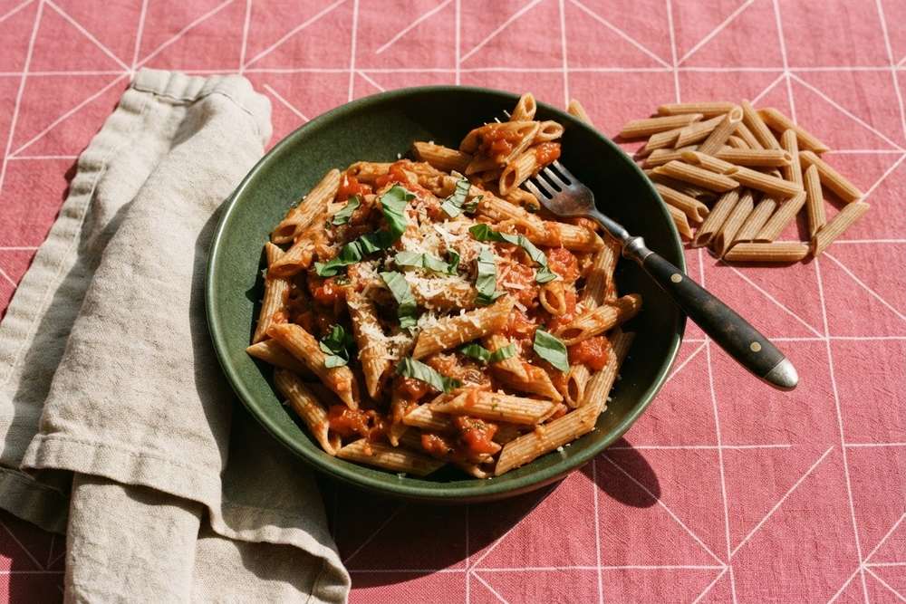 Tigela verde de macarrão integral ao sugo com manjericão e queijo ralado ao lado de penne cru, sobre toalha rosa