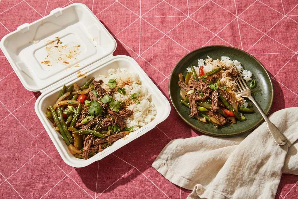Marmita de isopor aberta com arroz, carne desfiada e legumes ao lado do prato de cerâmica para onde a comida foi transferida, sobre toalha rosa