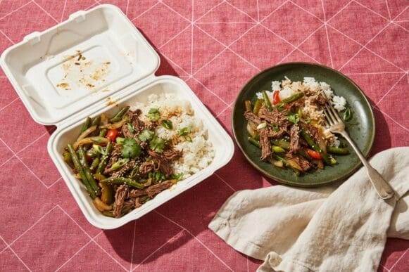 Marmita de isopor aberta com arroz, carne desfiada e legumes ao lado do prato de cerâmica para onde a comida foi transferida, sobre toalha rosa