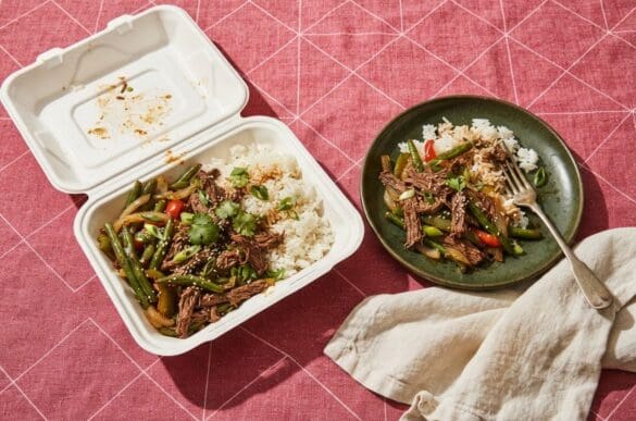 Marmita de isopor aberta com arroz, carne desfiada e legumes ao lado do prato de cerâmica para onde a comida foi transferida, sobre toalha rosa