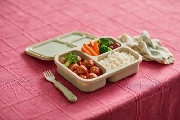 Marmita infantil com almôndegas ao molho, arroz, cenoura e brócolis em recipiente com divisórias sobre toalha rosa