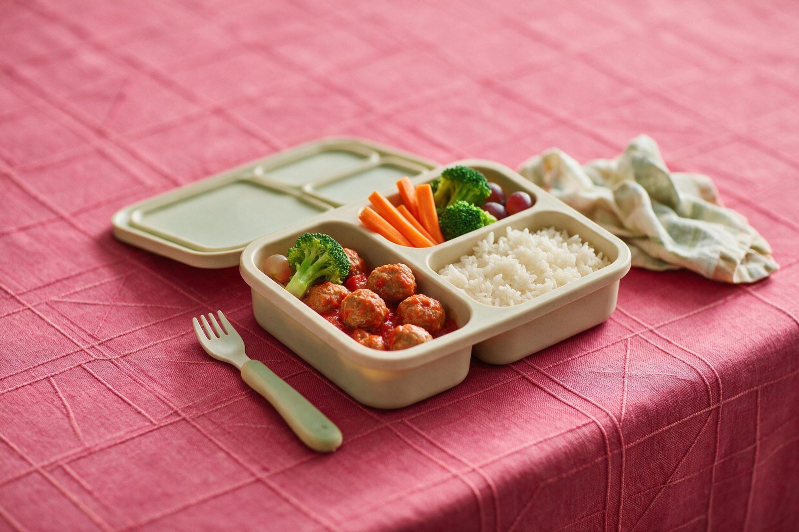 Marmita infantil com almôndegas ao molho, arroz, cenoura e brócolis em recipiente com divisórias sobre toalha rosa