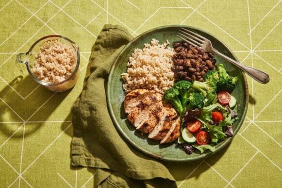 Prato verde com frango grelhado, arroz integral, feijão e brócolis ao lado de um copo de água, sobre toalha verde-limão
