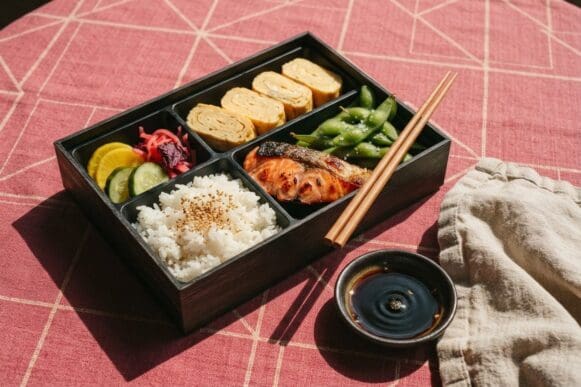 Bentô com arroz, salmão grelhado, omelete tamagoyaki, edamame e conservas, com hashi apoiado, sobre toalha rosa