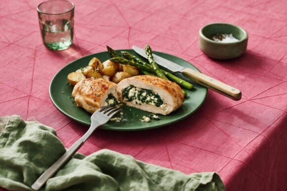 Peito de frango recheado com espinafre e queijo branco, fatiado, com batatas assadas e aspargos em prato verde sobre toalha rosa