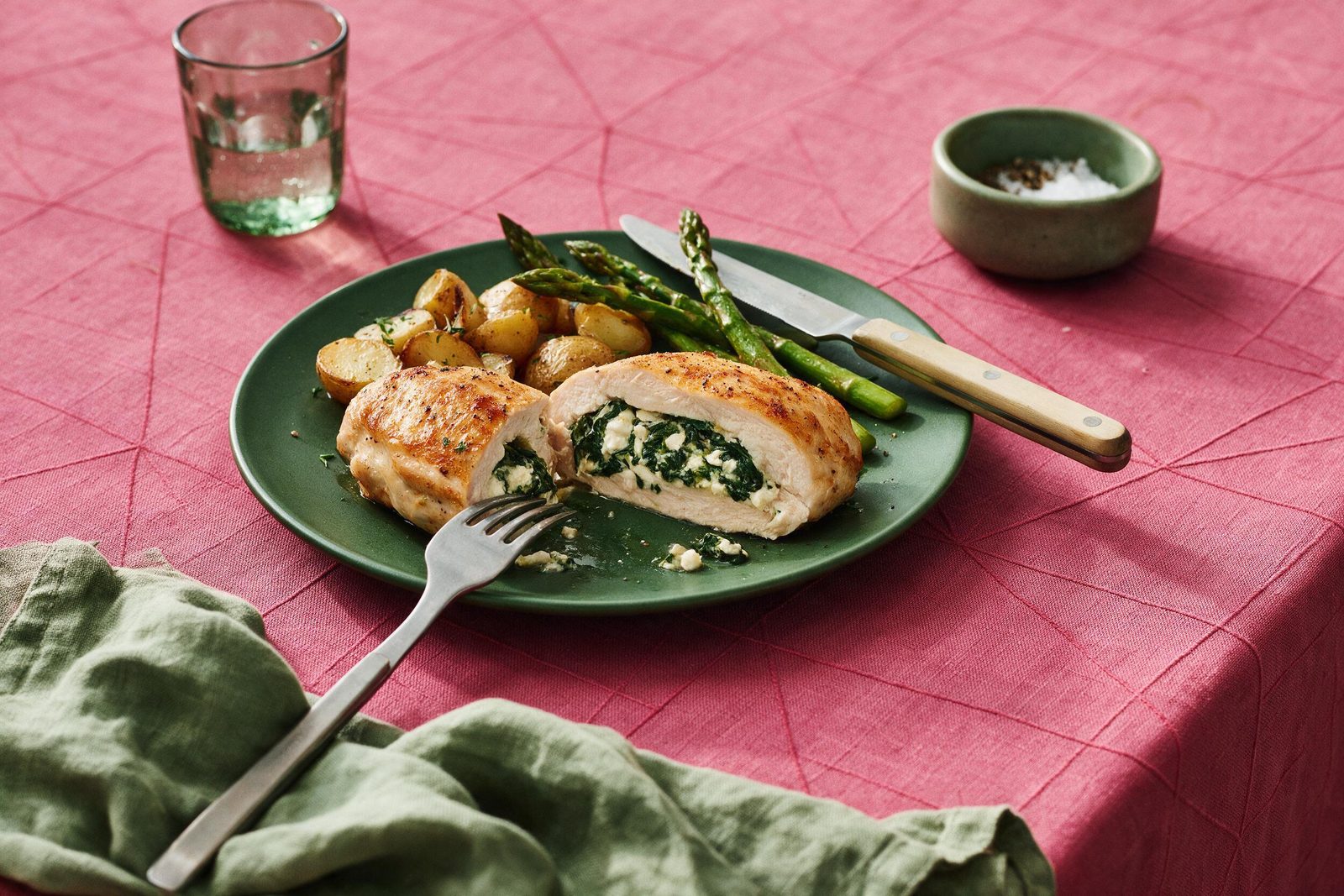 Peito de frango recheado com espinafre e queijo branco, fatiado, com batatas assadas e aspargos em prato verde sobre toalha rosa