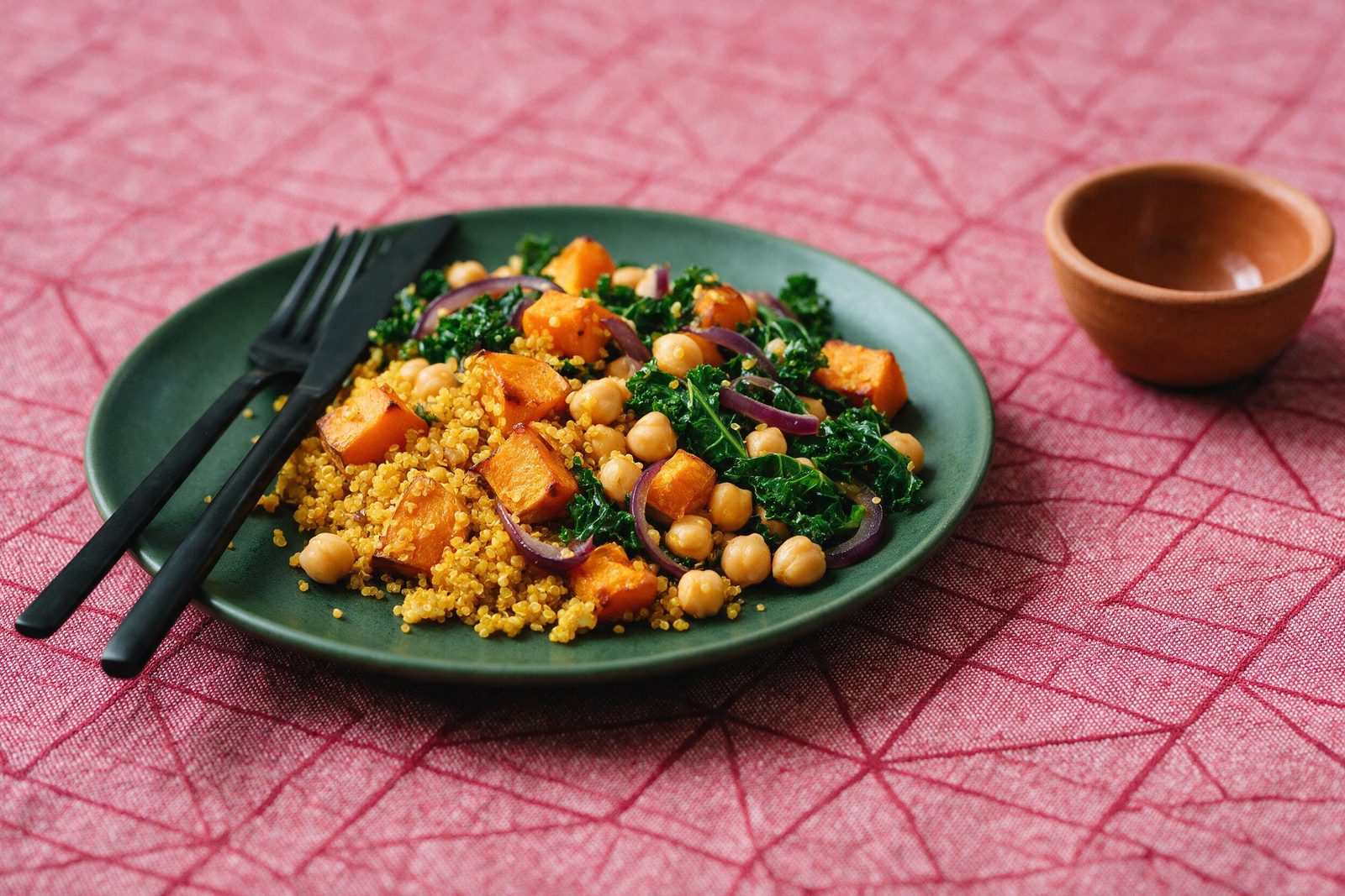 Prato verde com quinoa, abóbora assada, grão-de-bico e couve refogada sobre toalha rosa