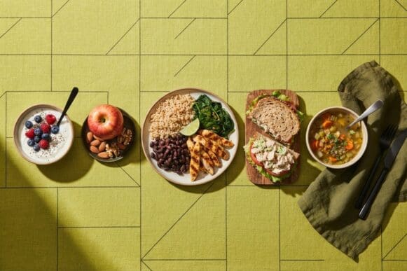 As refeições do dia dispostas em cinco pratos: iogurte com frutas, fruta com castanhas, almoço completo, sanduíche e sopa, sobre toalha verde-limão