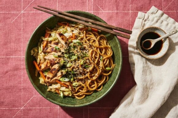 Tigela verde de yakissoba de frango com repolho, cenoura e gergelim, com hashi apoiado, sobre toalha rosa