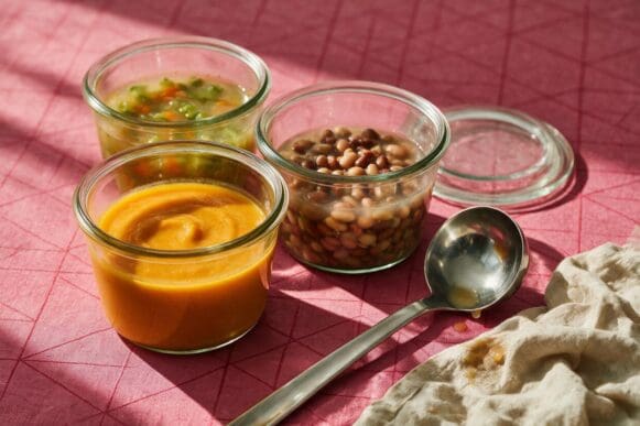 Potes de vidro com sopas de abóbora, legumes e feijão prontas para congelar, concha ao lado, sobre toalha rosa