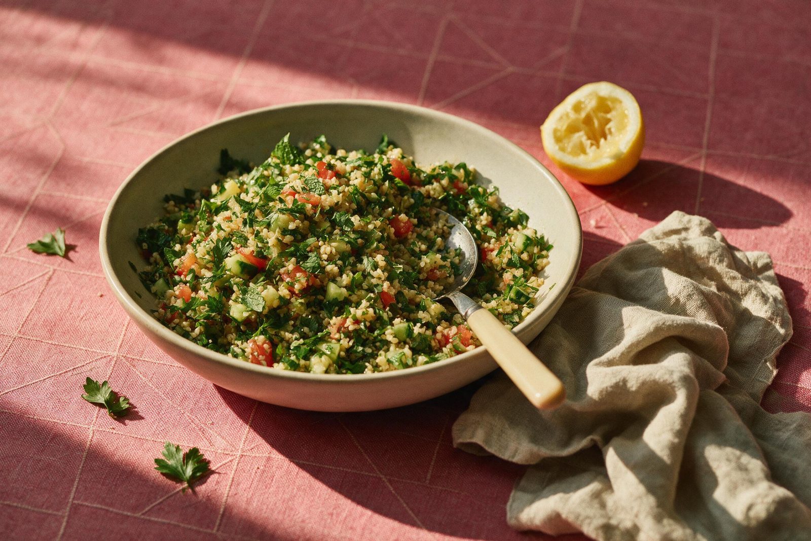 Travessa de tabule com trigo, salsinha, tomate, pepino e hortelã, meio limão espremido ao lado, sobre toalha rosa